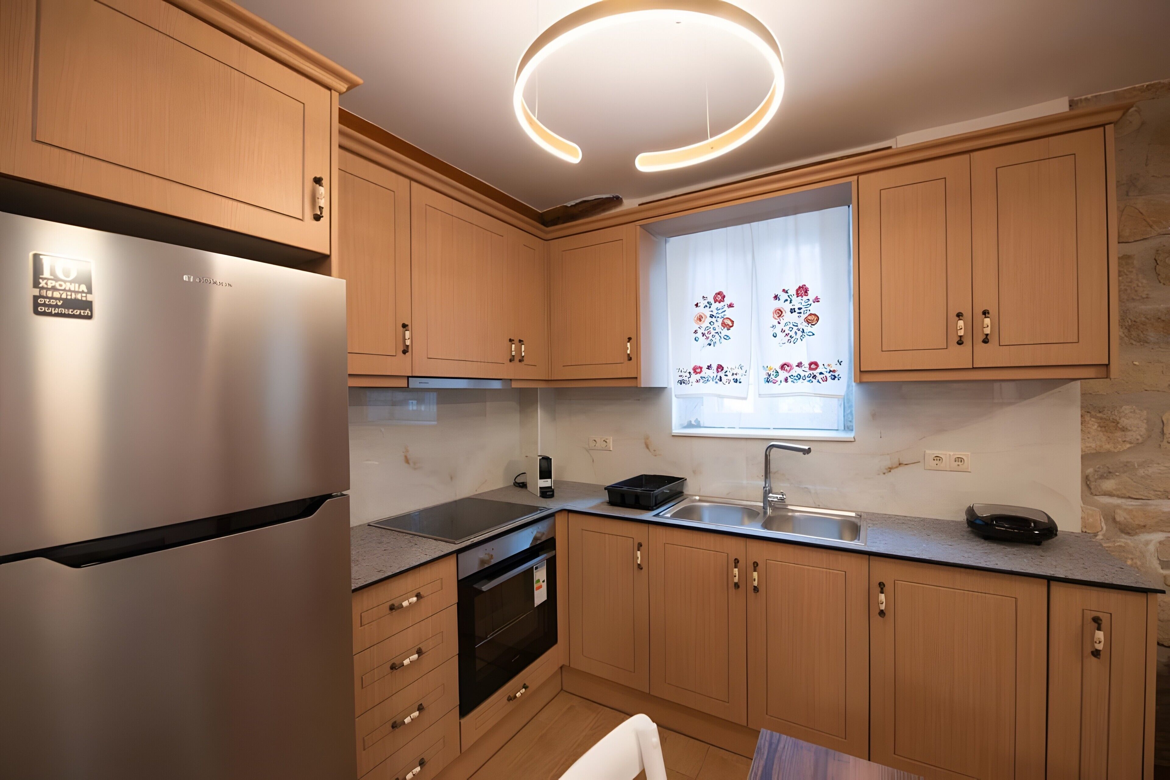 A modern kitchen with wooden cabinets, a stainless steel refrigerator, a built-in oven, and a window with decorative curtains.