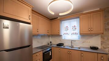 A modern kitchen with wooden cabinets, a stainless steel refrigerator, a built-in oven, and a window with decorative curtains.
