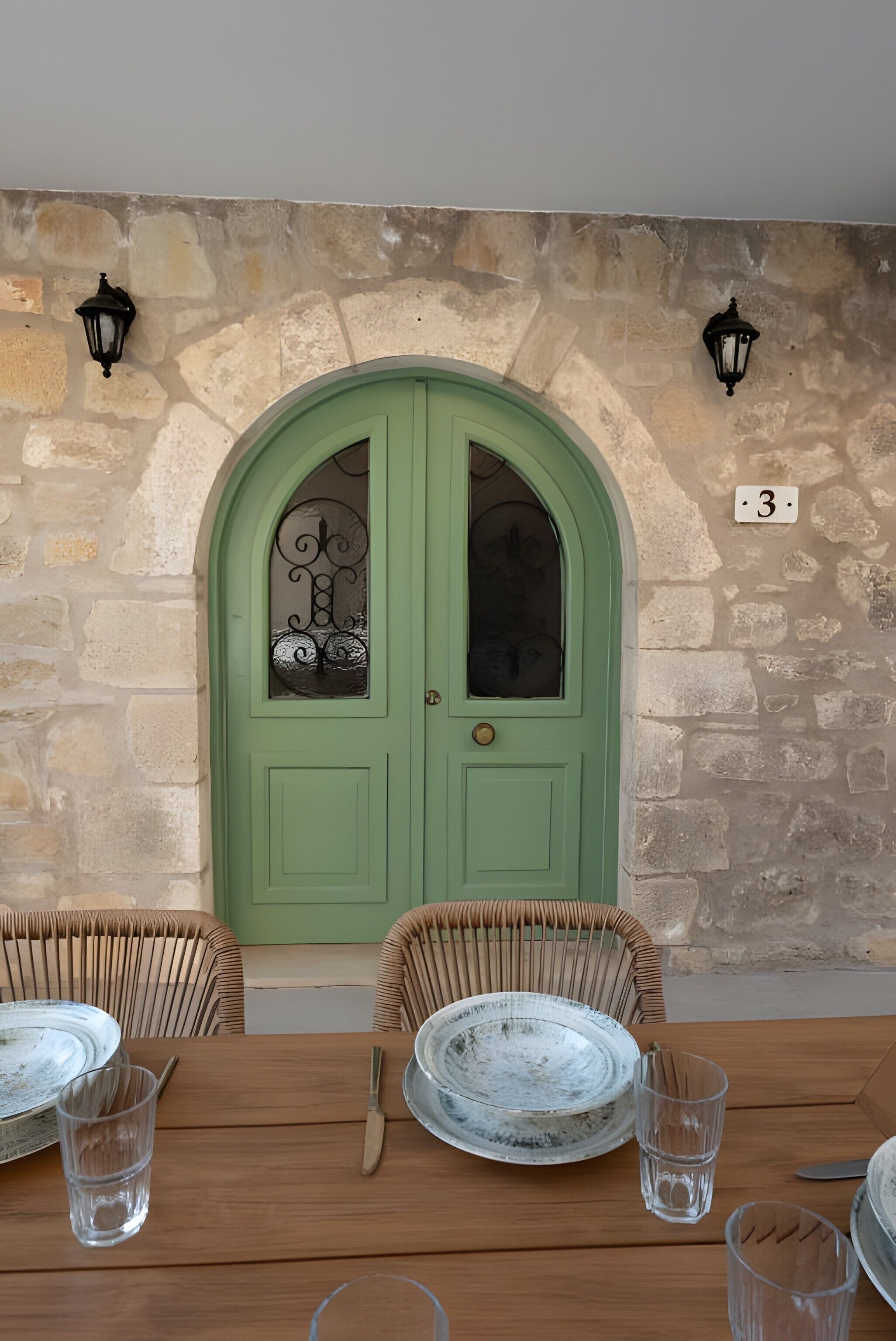 Uma porta verde com painel de vidro decorativo, parede de pedra e duas luzes fixadas na parede.