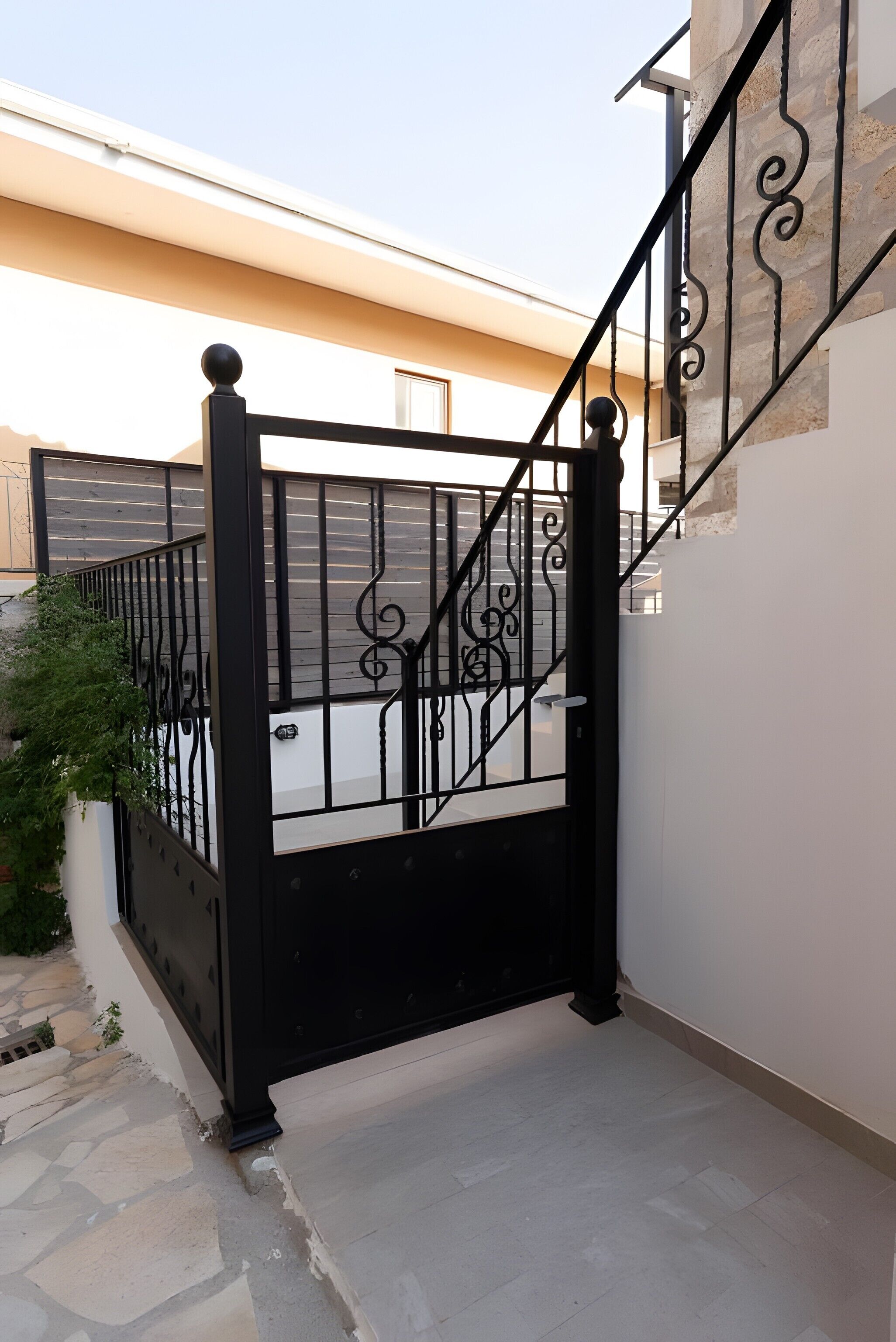 A black metal gate with intricate designs, leading to a staircase with a stone wall on one side and a beige wall on the other.
