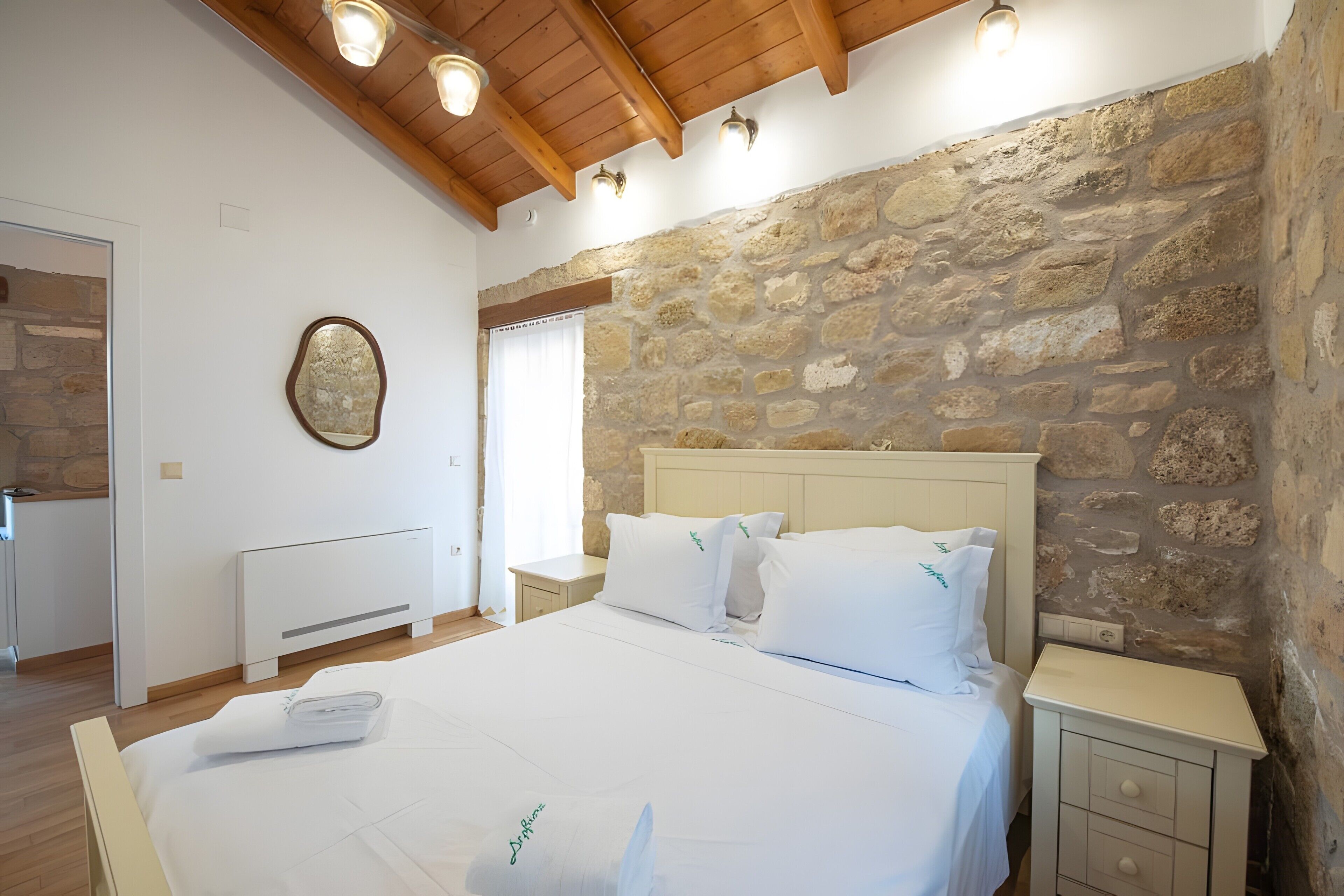 Ein Schlafzimmer mit Steinwand, Holzboden und einem Bett mit weißer Bettwäsche, sowie einem Nachttisch mit Lampe.
