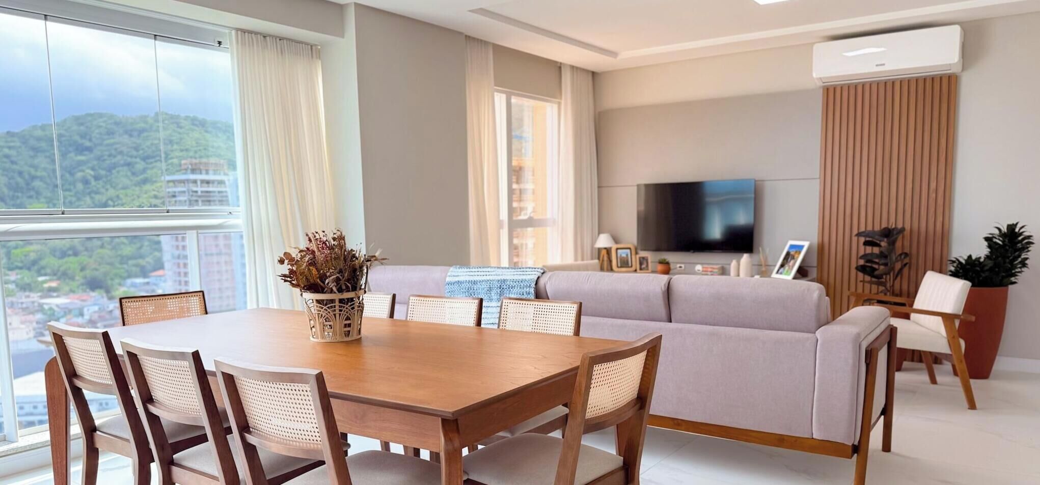 Uma sala de estar moderna com área de jantar, um conjunto de sofás, televisão de tela plana e uma grande janela com vista para o horizonte urbano.