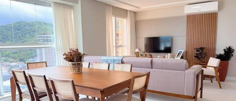Una sala moderna con comedor, un conjunto de sofás, un televisor de pantalla plana y un ventanal con vista a un paisaje urbano.