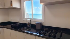 A kitchen with a double sink, a gas stove, and a window overlooking the sea. - ASA Vista Mar 3QT Alto Padrao Itapema (Itapema)
