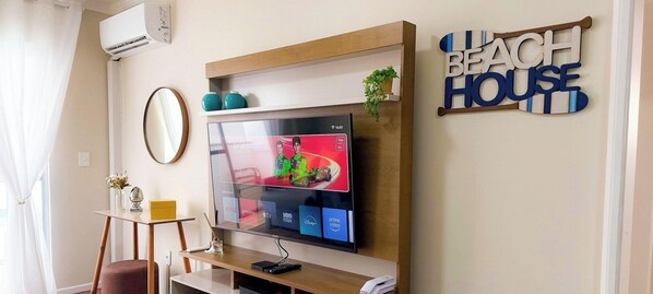 A living room with a flat-screen TV mounted on a wooden shelf, a small table with decorative items, a mirror, and a wall sign that reads 'BEACH HOUSE'. - ASA 3QT Leblon Ar Av. Brasil Centro BC (Balneário Camboriú)