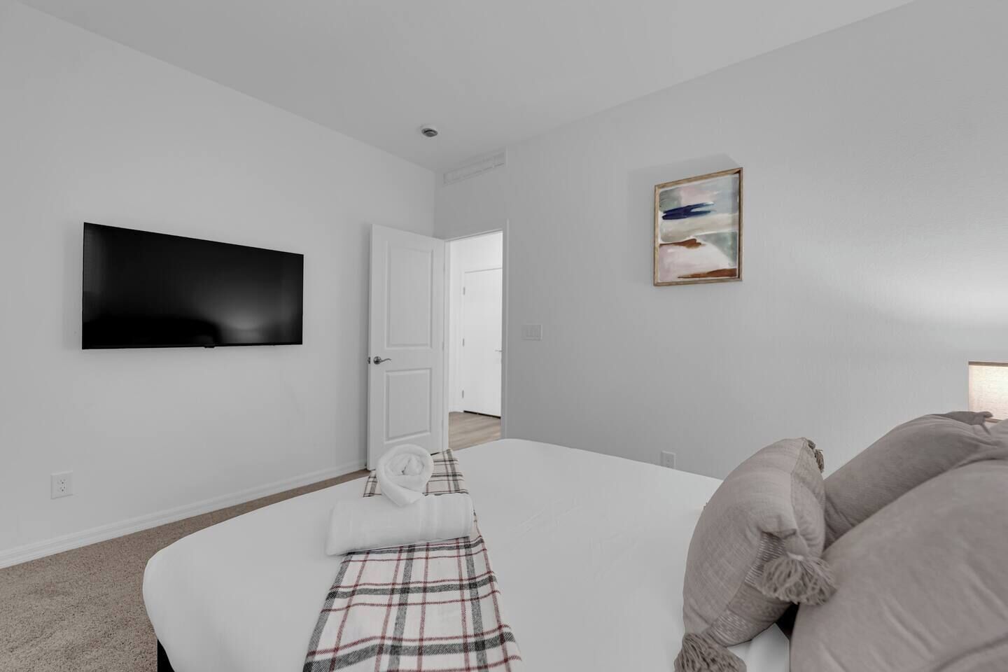A modern hotel room with a flat-screen TV mounted on the wall, a bed with a plaid blanket, a door leading to another room, and a painting on the wall.