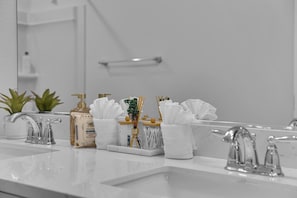Een badkamer met een witte wastafel, twee kranen, een spiegel en diverse toiletartikelen, waaronder een fles Brazilian mist, een verpakking 3D-doekjes en een vaas met een plant.