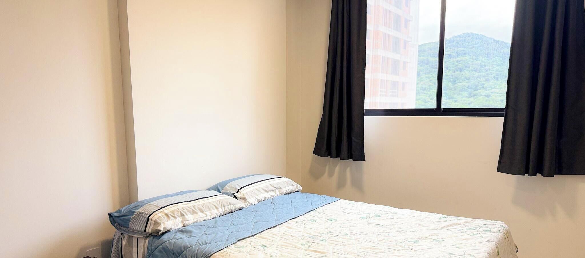 A single bed with a blue and white duvet, two pillows, and a window with black curtains.