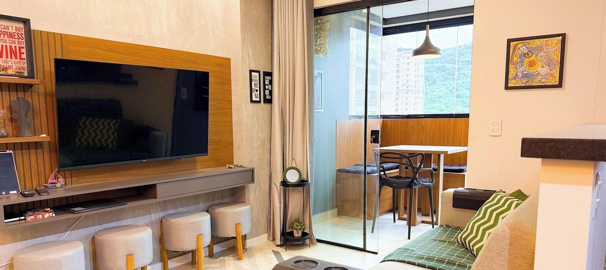 A modern living room with a large flat-screen TV, a bar area with stools, a dining table with chairs, and a sliding glass door leading to a balcony.