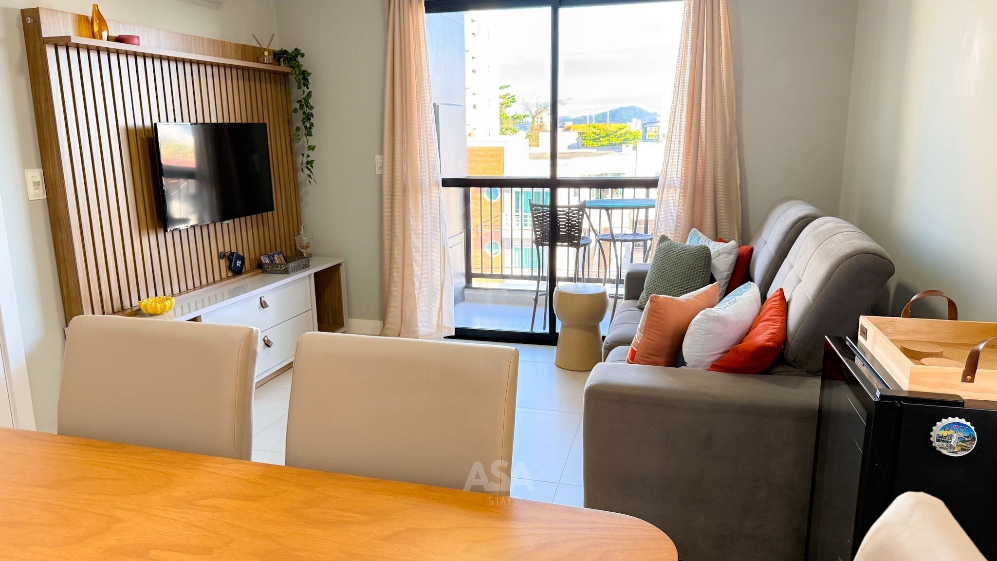 A modern living room with a flat-screen TV, a sofa with cushions, a dining table, and a balcony with outdoor furniture.