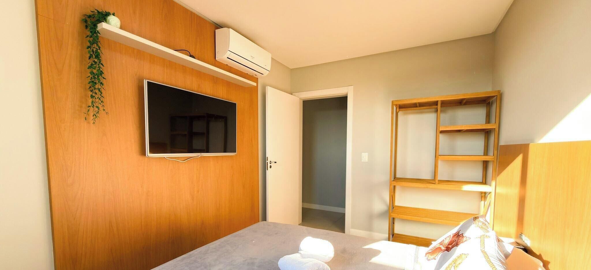 A room with a wooden wardrobe, a mounted TV, a shelf with a plant, and a door leading to another room.