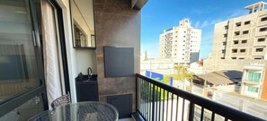 Varanda com mesa de vidro e uma cadeira, pia pequena e vista para edifícios ao redor.