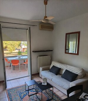 Een woonkamer met een bank, een klein tafeltje, een plafondventilator en uitzicht door een glazen deur op een buitenruimte met stoelen en een tafel.