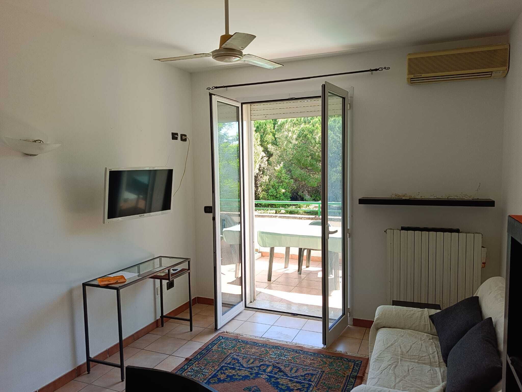 Ein Zimmer mit Deckenventilator, Fernseher, Tisch mit Glasplatte, einer Couch und Blick auf eine Außenpatio.
