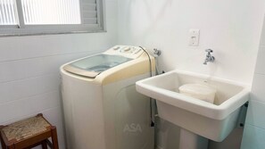 A compact laundry room with a washing machine, a sink, and a small wooden stool. - ASA Apto Central 2 QT Climatizado BC (Balneário Camboriú)