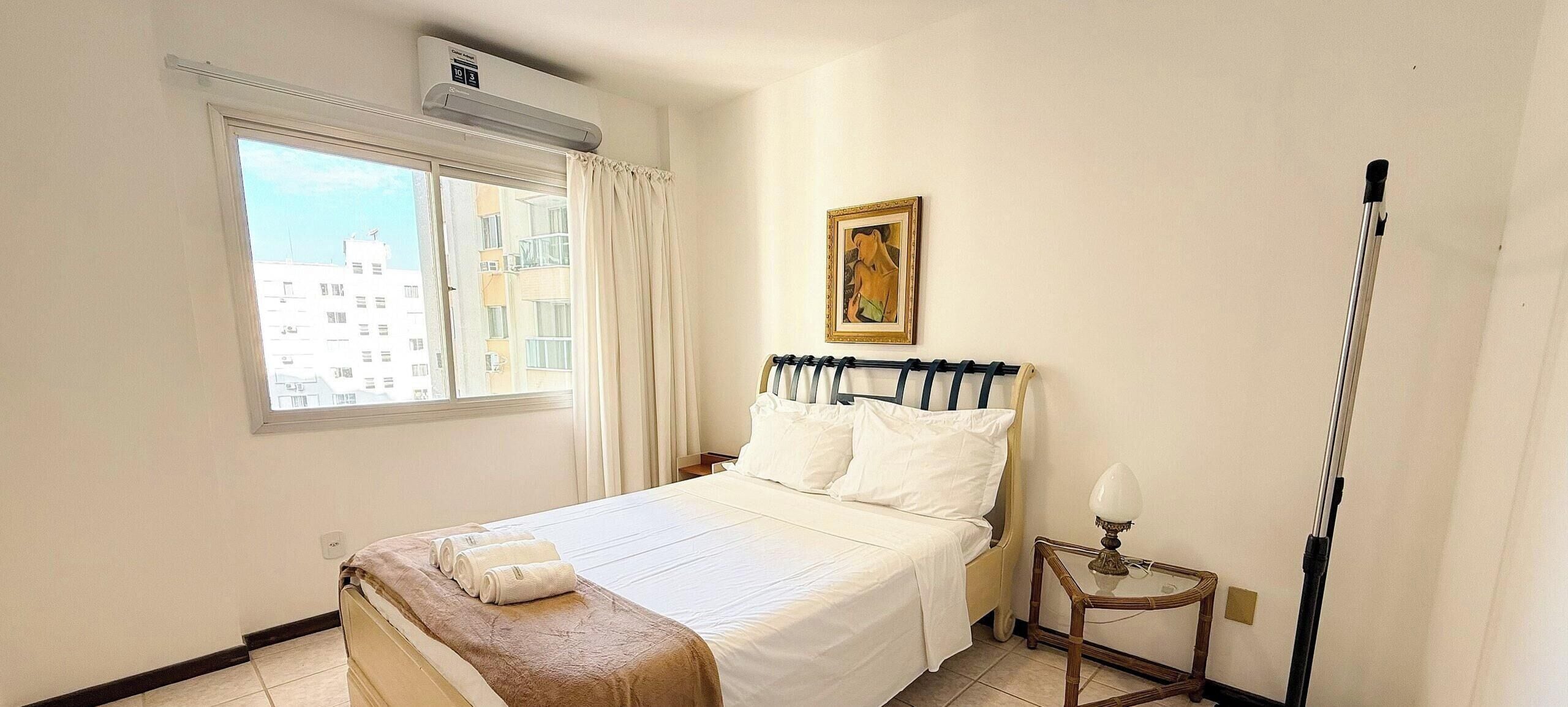 A hotel room with a bed, a window, a painting, and a bedside table.