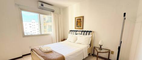 A hotel room with a bed, a window, a painting, and a bedside table.