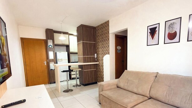 Una sala de estar con un sofá color crema, un televisor, una cocina con refrigerador y una mesa de comedor con sillas.