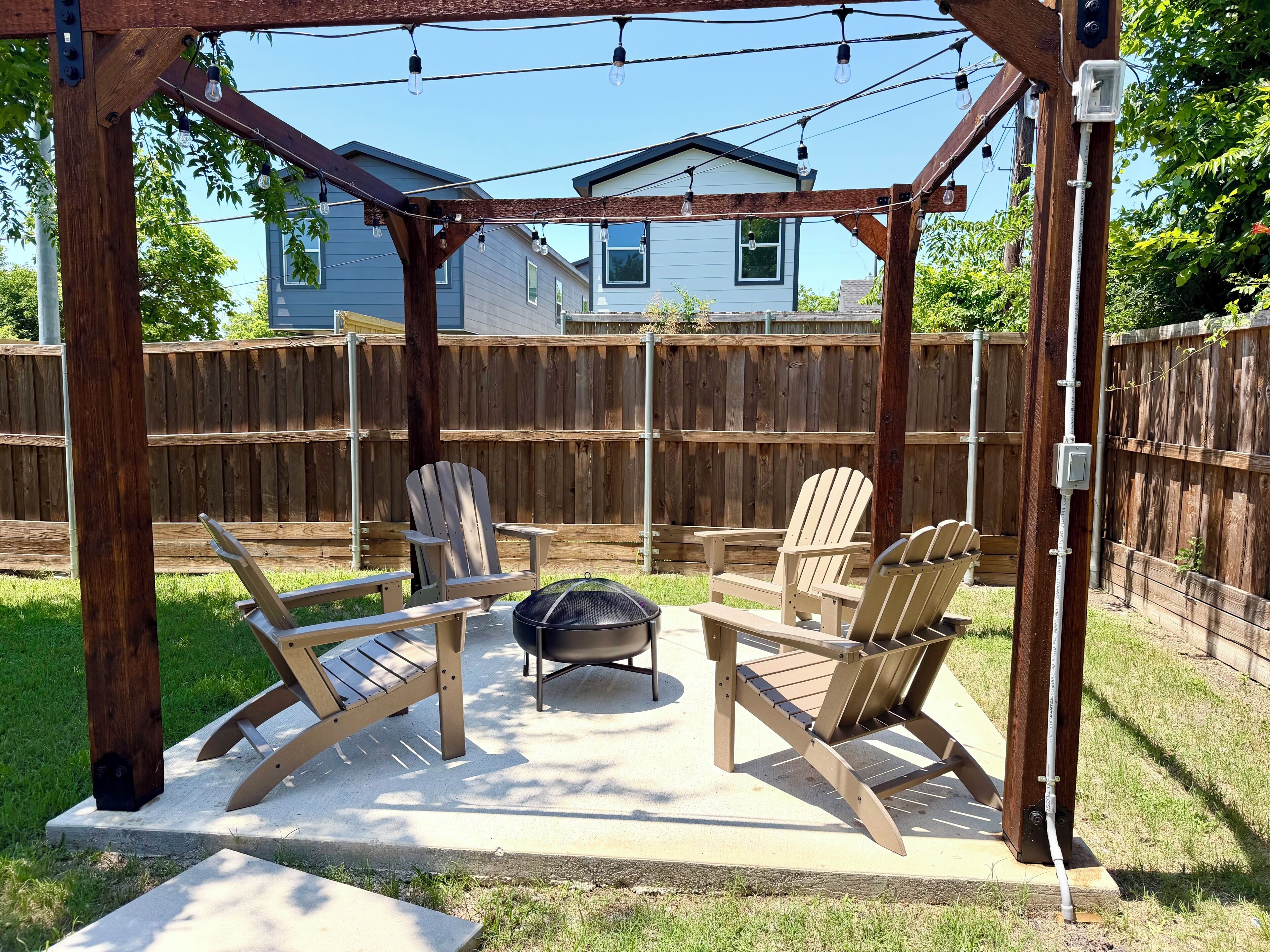 A covered patio with Adirondack chairs and a grill, surrounded by a wooden fence and houses in the background.