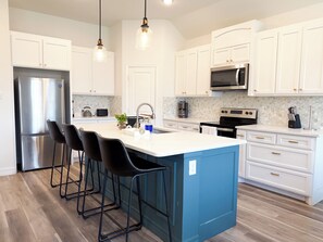 A modern kitchen with white cabinets, stainless steel appliances, a blue accent wall, and bar stools. - Modern 4BR Home • 5 Min Downtown & Fair Park (Dallas)