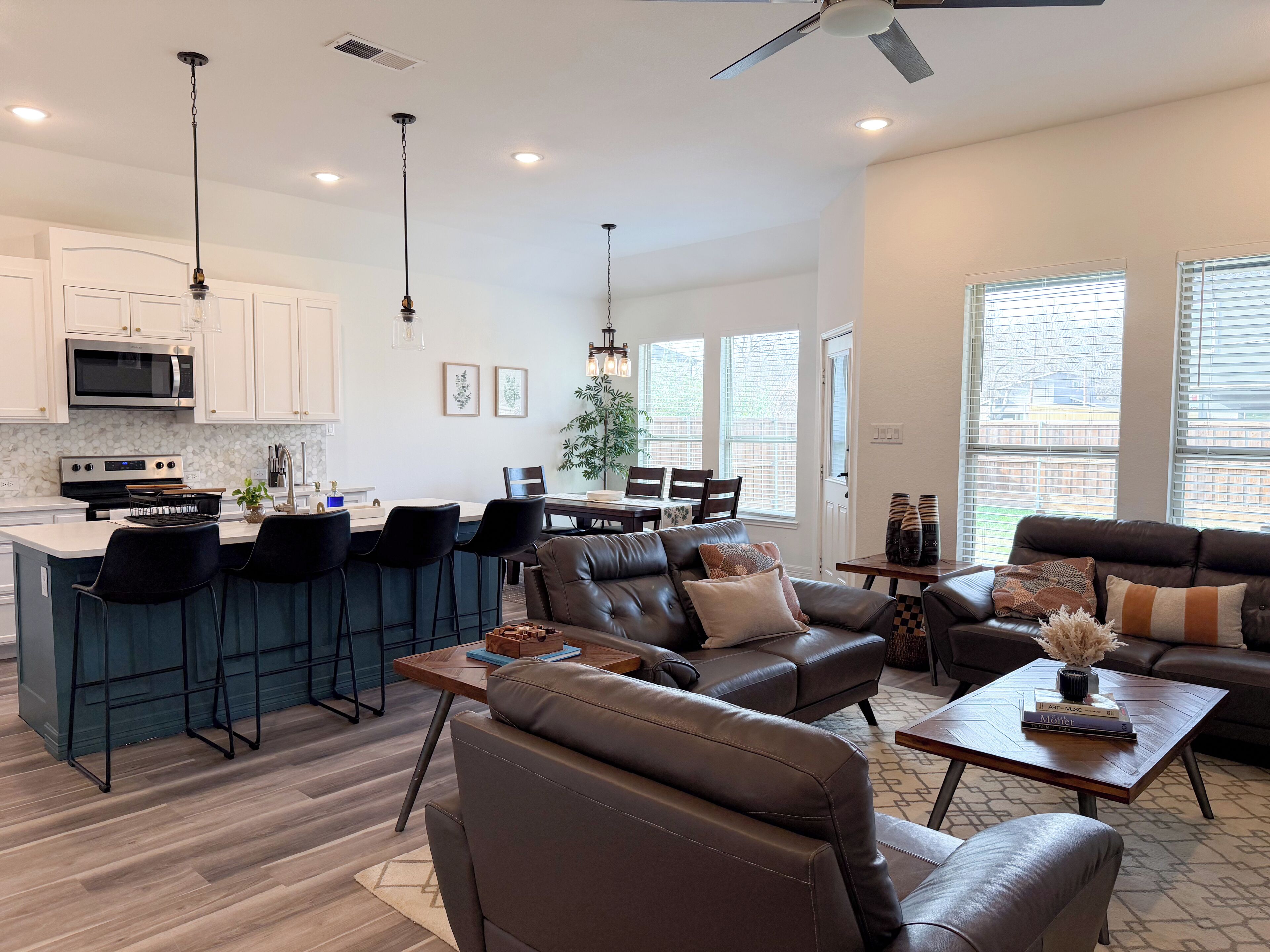 A modern kitchen with white cabinets, a microwave, and a stove. There is a dining area with a table and chairs, and a living room with a leather sofa and a coffee table.