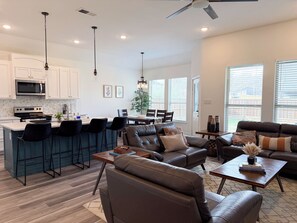 A modern kitchen with white cabinets, a microwave, and a stove. There is a dining area with a table and chairs, and a living room with a leather sofa and a coffee table. - Modern 4BR Home • 5 Min Downtown & Fair Park (Dallas)