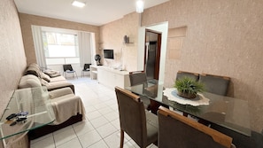 Uma sala de estar moderna com uma mesa de jantar de vidro, um conjunto de sofá, uma televisão e uma área de cozinha com geladeira.