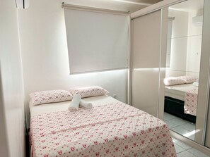 A bedroom with a bed, pillows, a bedside table, a mirror, and a window with a blind. - ASA Apto Funcional Prox. Praia Centro BC (Balneário Camboriú)