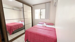 Hotellihuone, jossa on kaksi sänkyä, ja molemmissa on punavalkoruudullinen sängynpeite. Lisäksi huoneessa on suuri peili, ikkuna, jossa on verhot, ja ilmastointilaite.