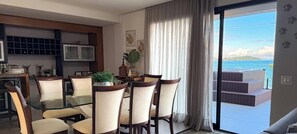 A dining area with a glass table and chairs, a kitchen with cabinets, and a view of the sea through a balcony. - ASA Beachfront 5 Suites Jacuzzi Bombinhas (Bombinhas)