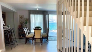 A dining area with a view of the sea, a staircase, and a living room with a large window. - ASA Beachfront 5 Suites Jacuzzi Bombinhas (Bombinhas)