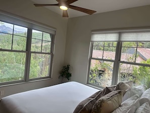 Ein Schlafzimmer mit einem Bett, zwei Fenstern, einem Deckenventilator und Blick auf einen Wald und Gebäude.