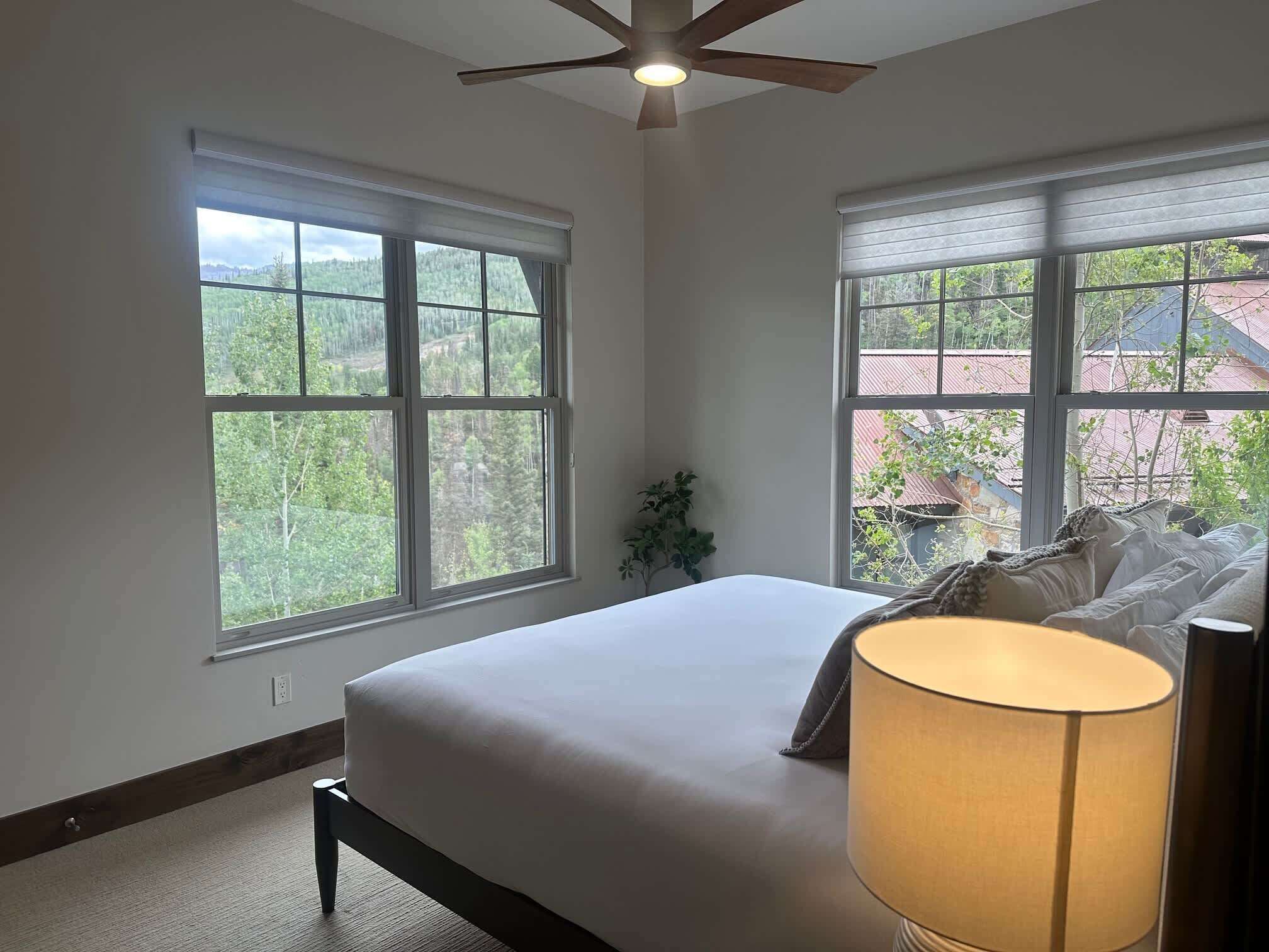 A bedroom with a bed, a ceiling fan, and two windows with a view of a forest and buildings.
