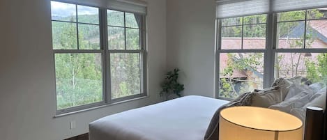 A bedroom with a bed, a ceiling fan, and two windows with a view of a forest and buildings.