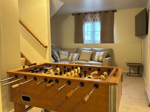 Ein Zimmer mit Tischfußball, einer Couch mit gemusterten Kissen, einem Fenster mit Vorhängen und einem an der Wand befestigten Fernseher.