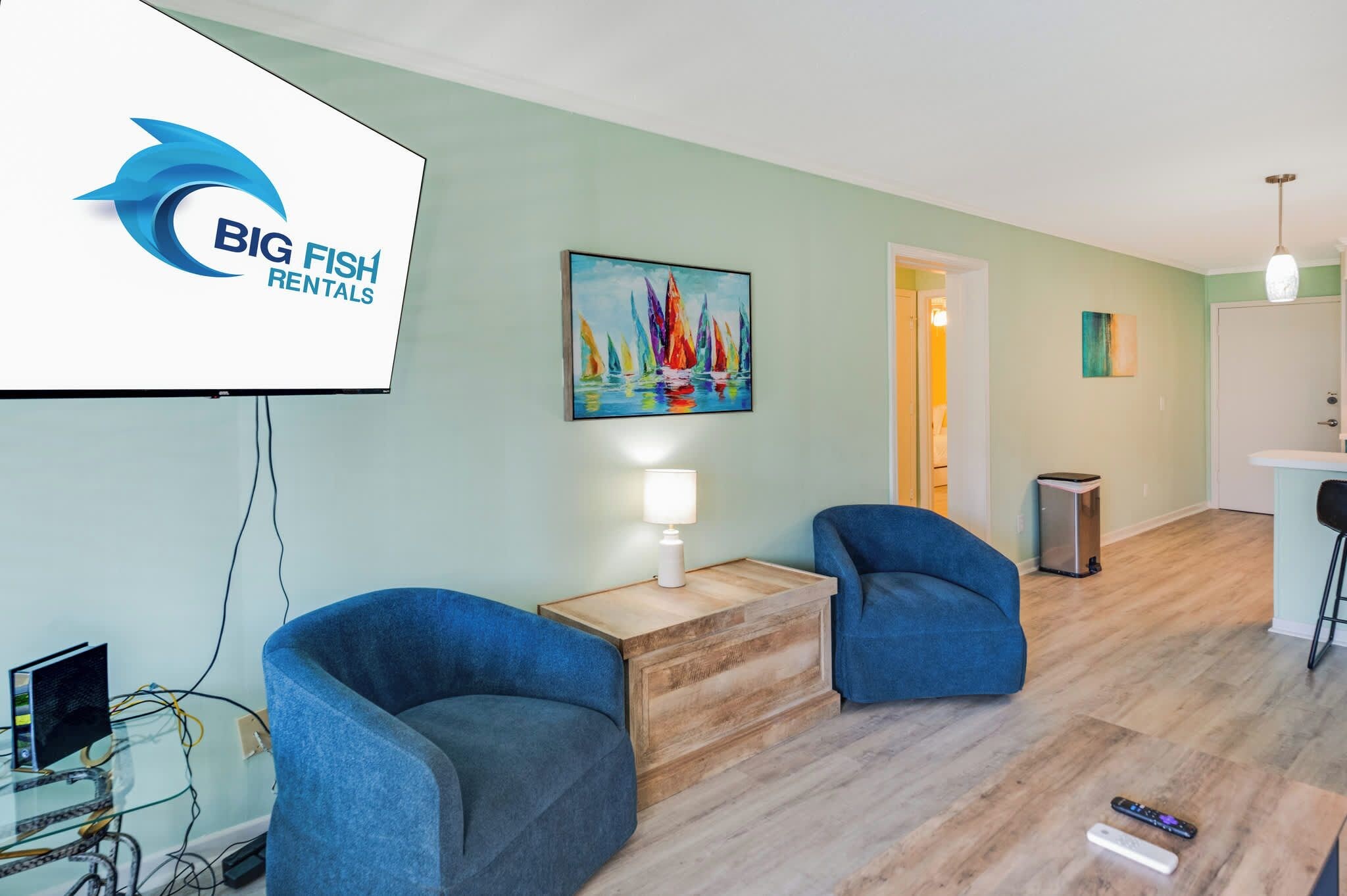Un salon moderne avec un téléviseur à écran plat, deux fauteuils bleus, une table basse en bois et un tableau coloré accroché au mur.