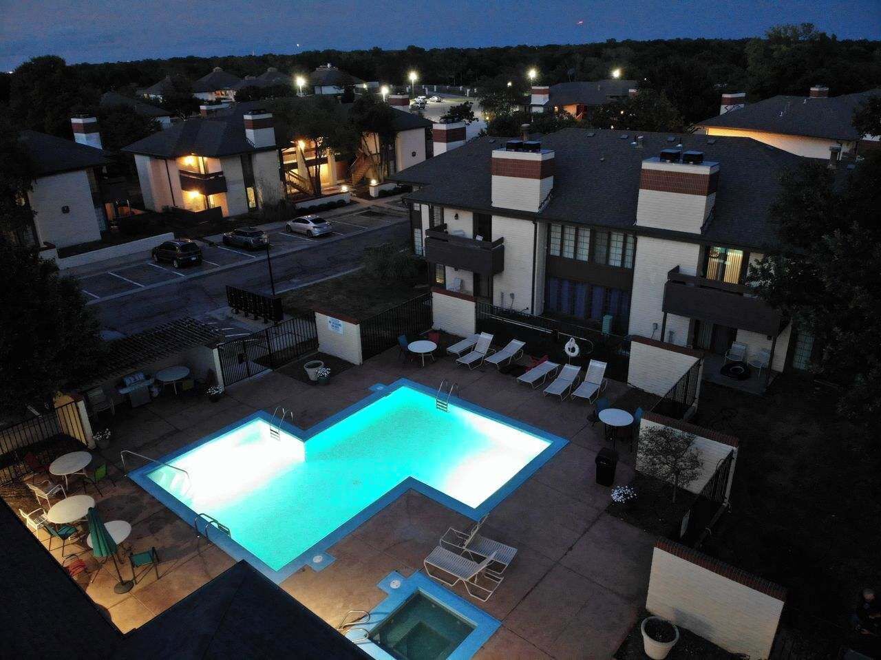 Une piscine entourée de bâtiments dont les fenêtres sont illuminées, avec des chaises longues et des tables à proximité.