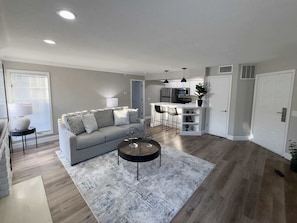 A modern living room with a grey sofa, a coffee table, and a kitchen area with a microwave and a refrigerator.