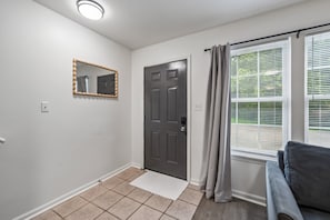 Une pièce avec une porte, un miroir, une fenêtre avec des stores et un petit tapis.
