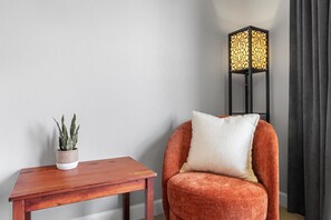 Ein Zimmer mit Holztisch, Topfpflanze, Sessel mit Kissen und Stehlampe.