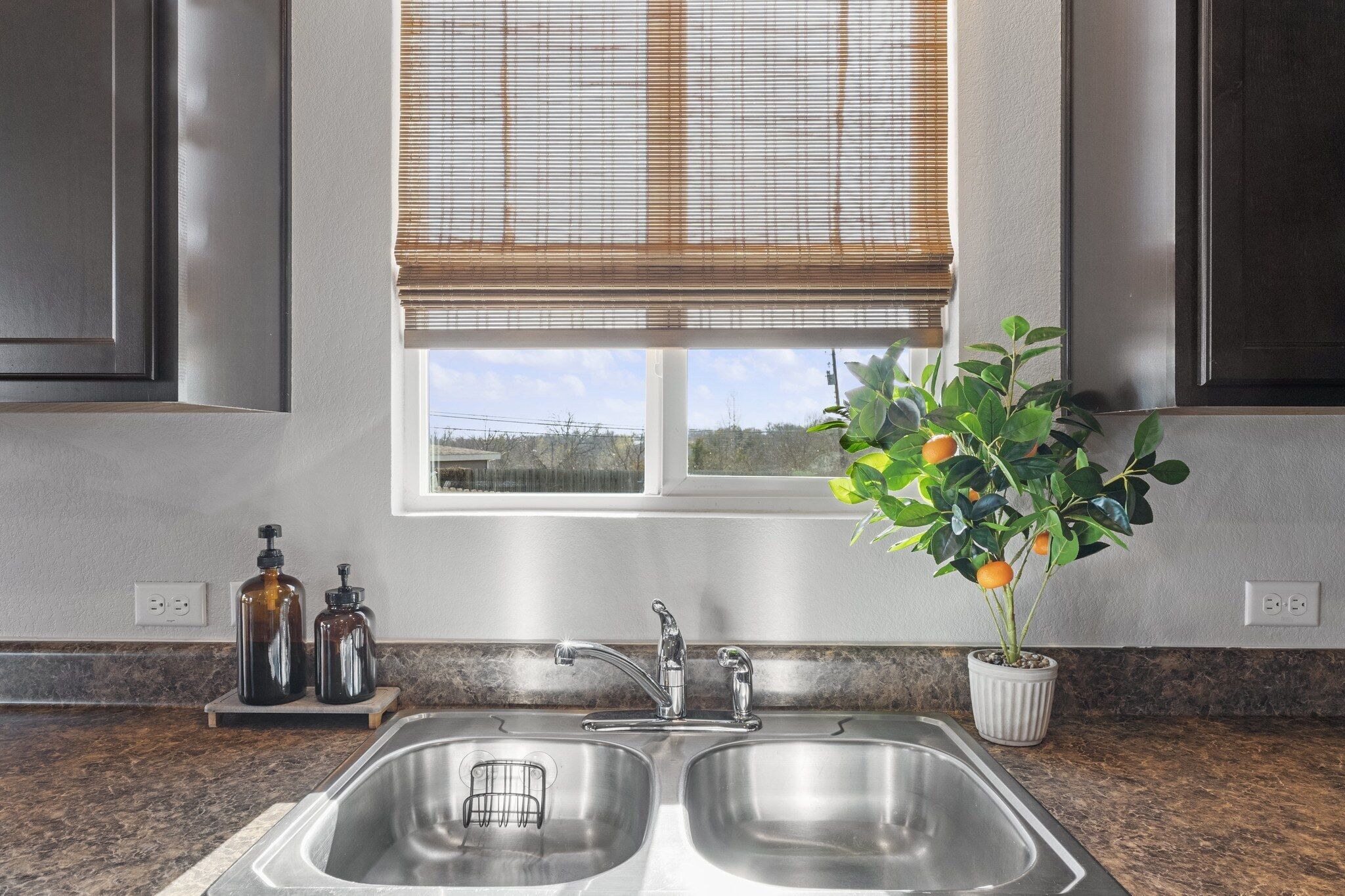 A kitchen with a double stainless steel sink, a window with blinds, and a potted plant with green leaves and orange fruits.