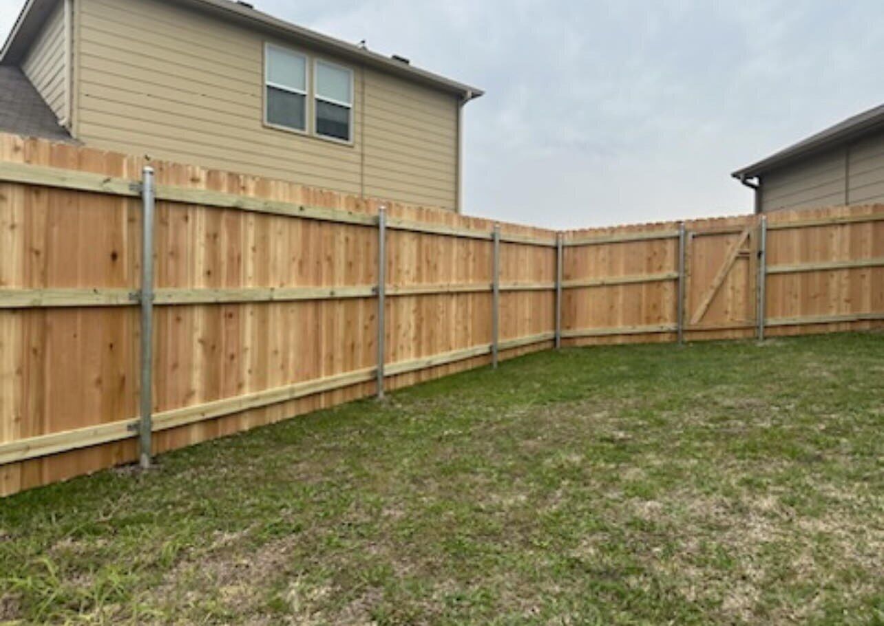 A fenced backyard with a wooden fence, a grassy lawn, and a house with a window.
