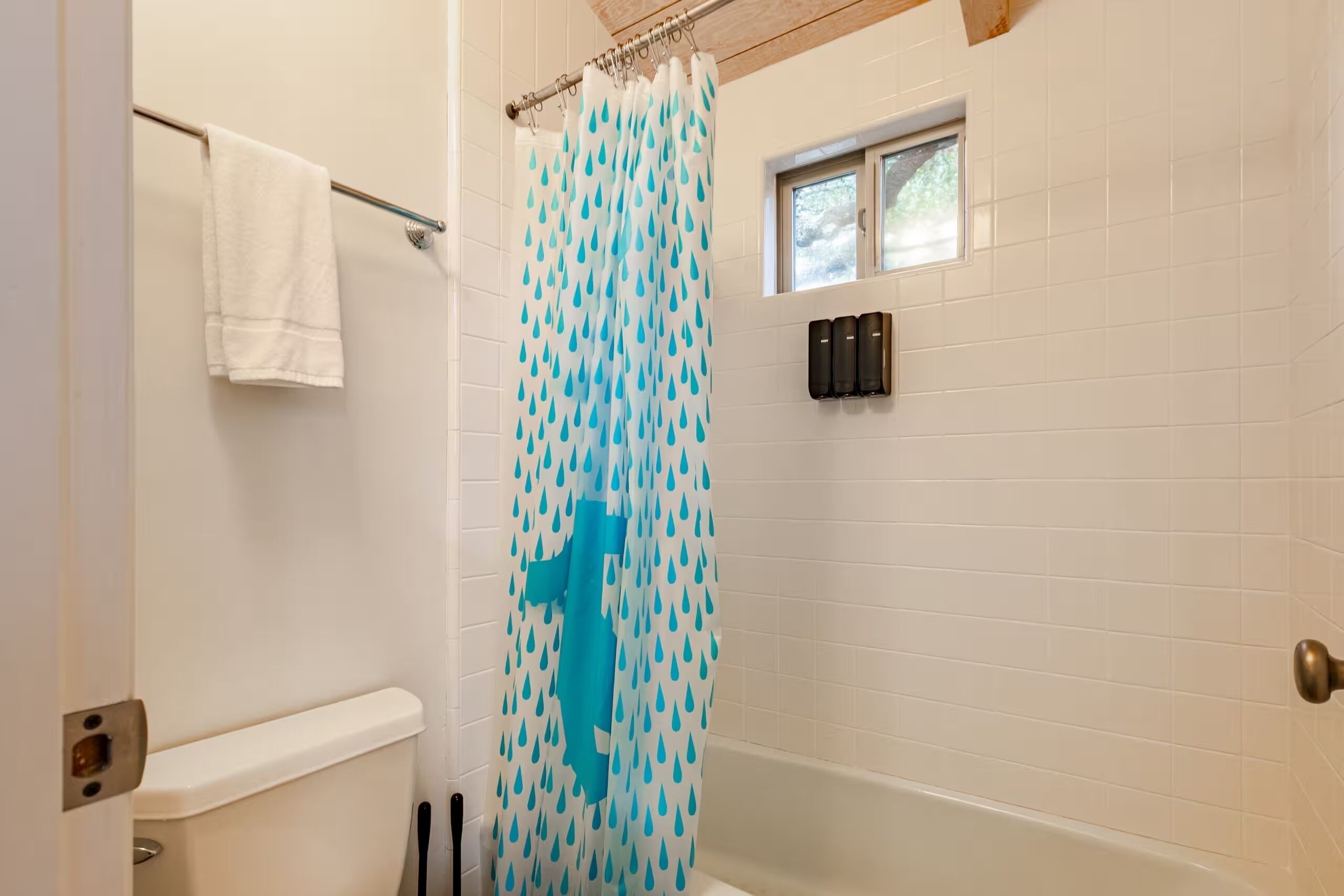Une salle de bain avec un mur recouvert de carreaux blancs, un rideau de douche à motifs bleus, une serviette blanche suspendue à une tringle métallique et une fenêtre située au-dessus de la douche.
