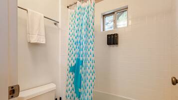 Une salle de bain avec un mur recouvert de carreaux blancs, un rideau de douche à motifs bleus, une serviette blanche suspendue à une tringle métallique et une fenêtre située au-dessus de la douche.