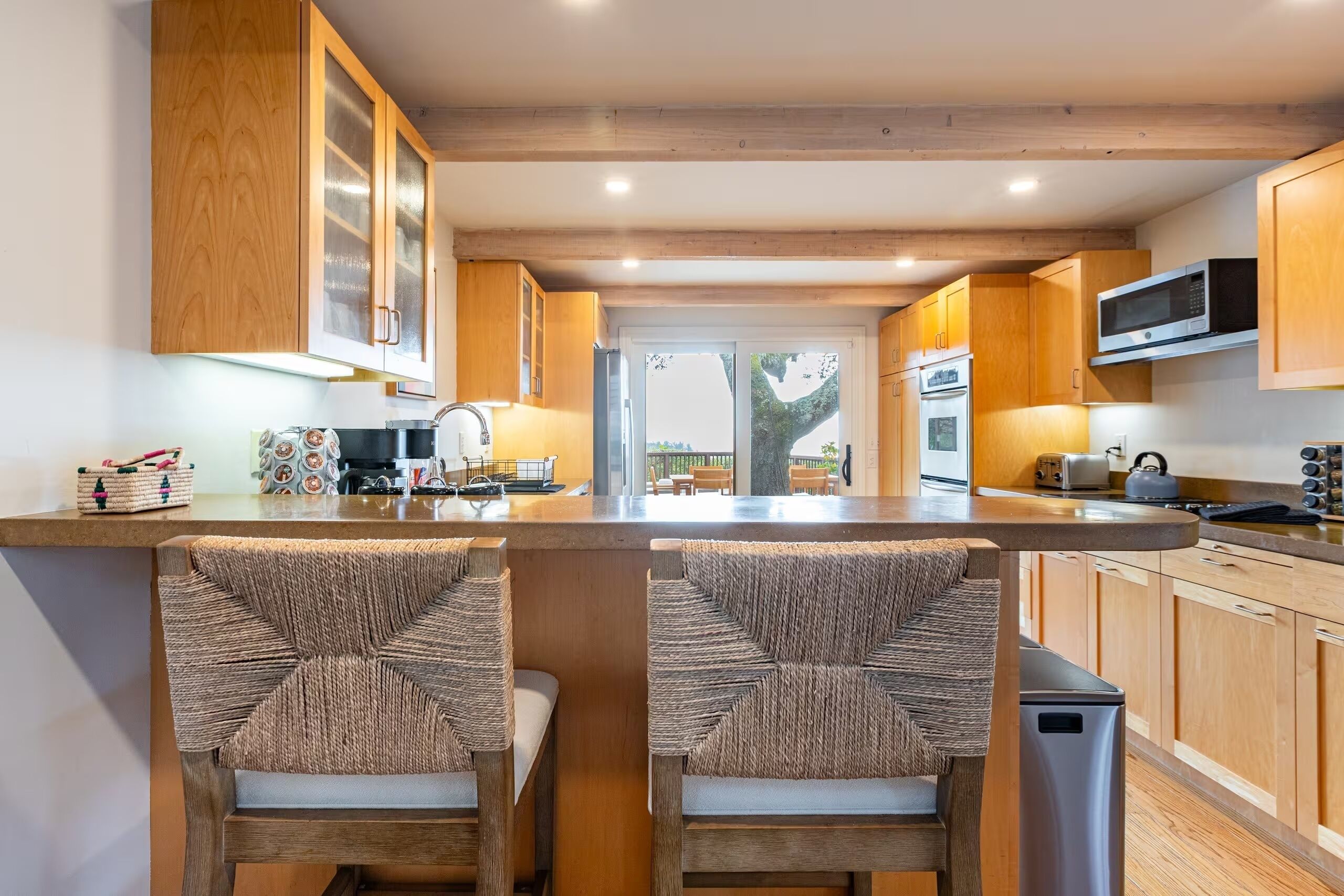 A modern kitchen with wooden cabinets, a central island with bar stools, stainless steel appliances, and a view through the door.