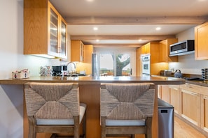 Une cuisine moderne avec des meubles en bois, un îlot central doté de tabourets de bar, des appareils en acier inoxydable et une vue sur l’extérieur par la porte.
