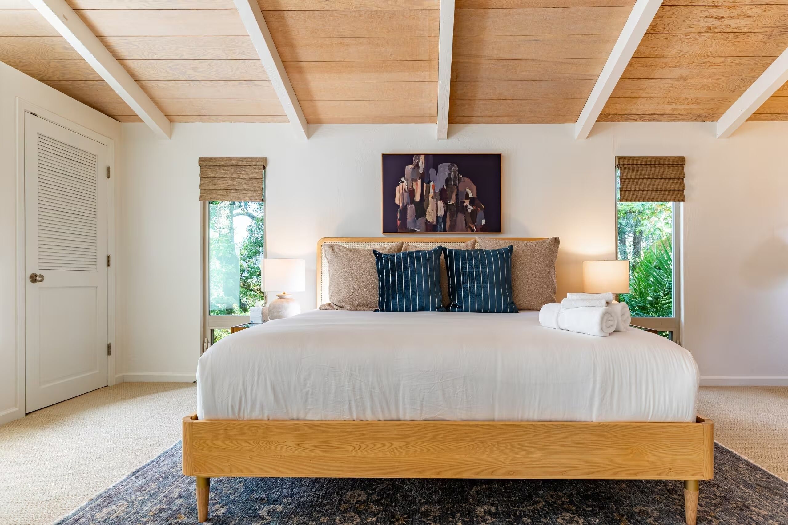 A bedroom with a wooden bed frame, a large window with a view of greenery, and a painting above the bed.