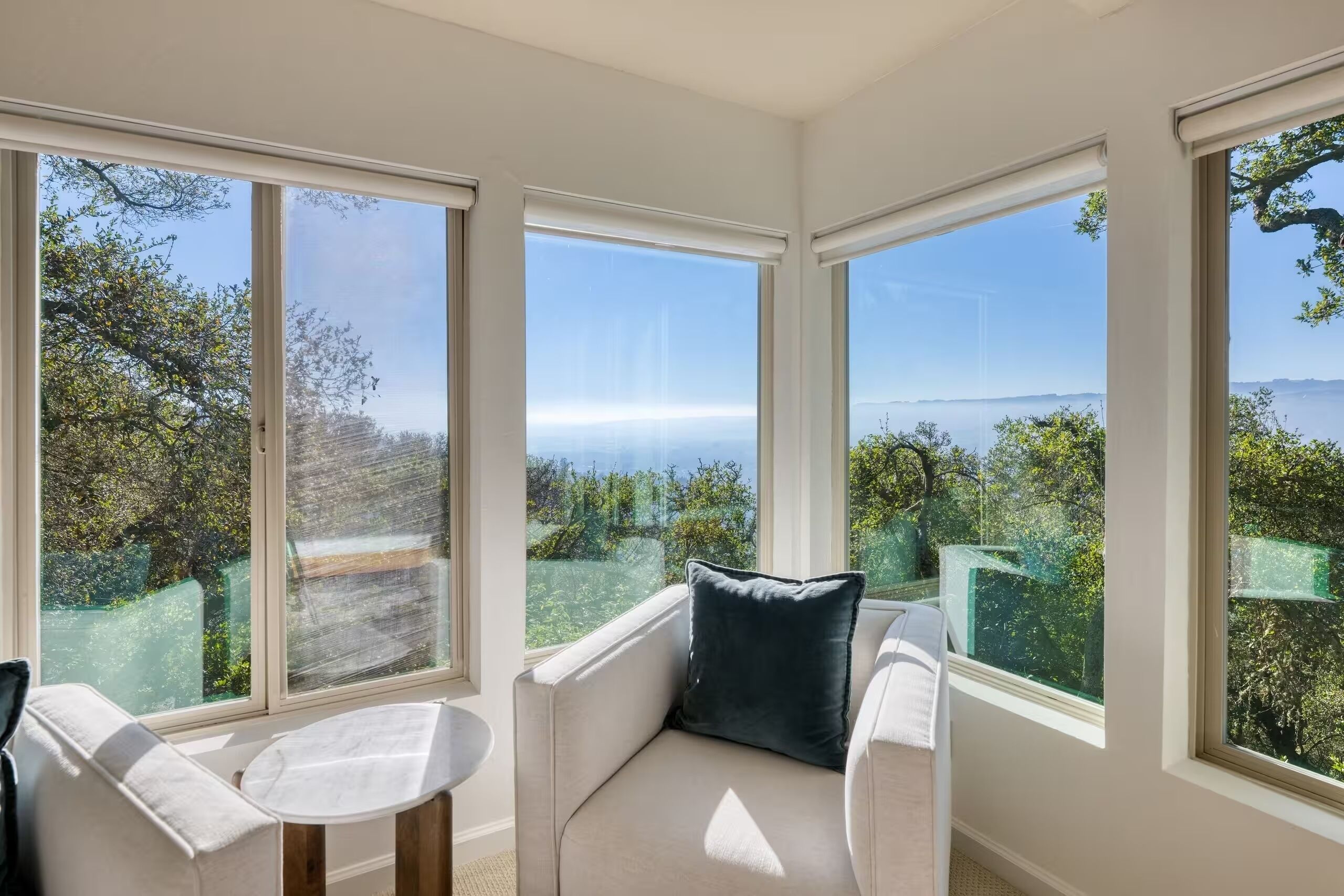 Una habitación con sofá y una pequeña mesa, ventanas amplias con vistas a árboles y un paisaje lejano.