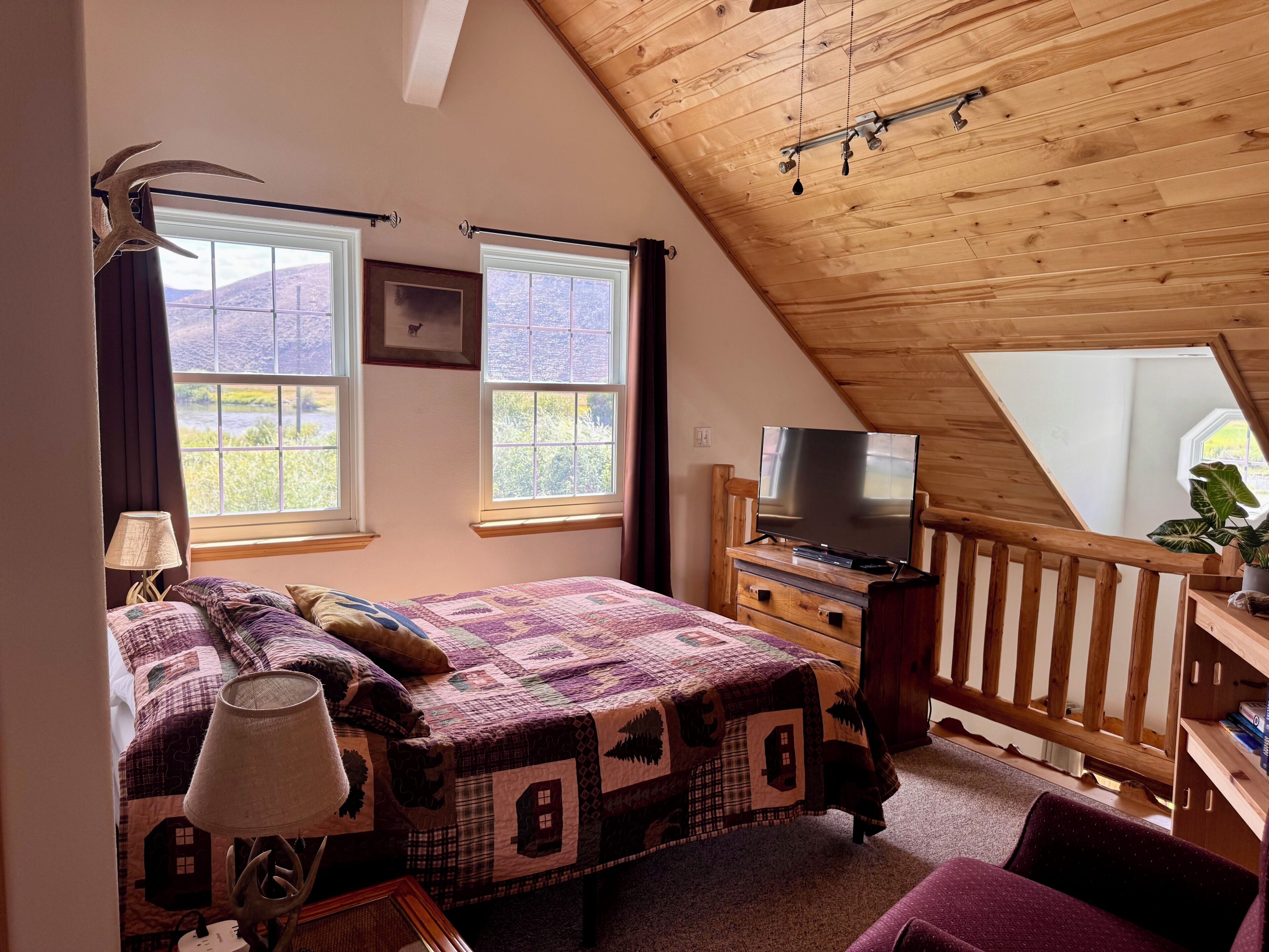 A bedroom with a sloped ceiling, a bed with a quilt, a television on a wooden dresser, and a window with a view of the outdoors.