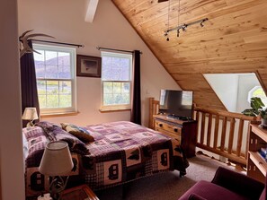A bedroom with a sloped ceiling, a bed with a quilt, a television on a wooden dresser, and a window with a view of the outdoors.